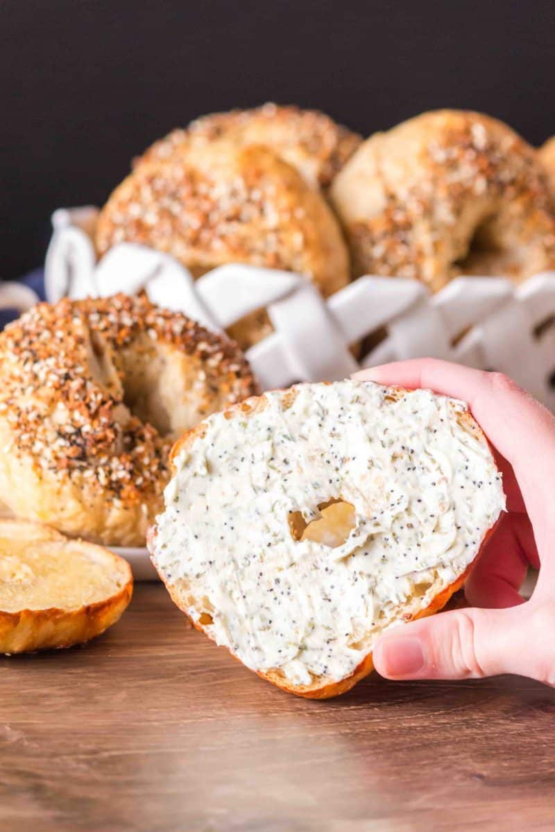 A hand holding a sliced half of a homemade bagel spread with cream cheese - Hostess At Heart