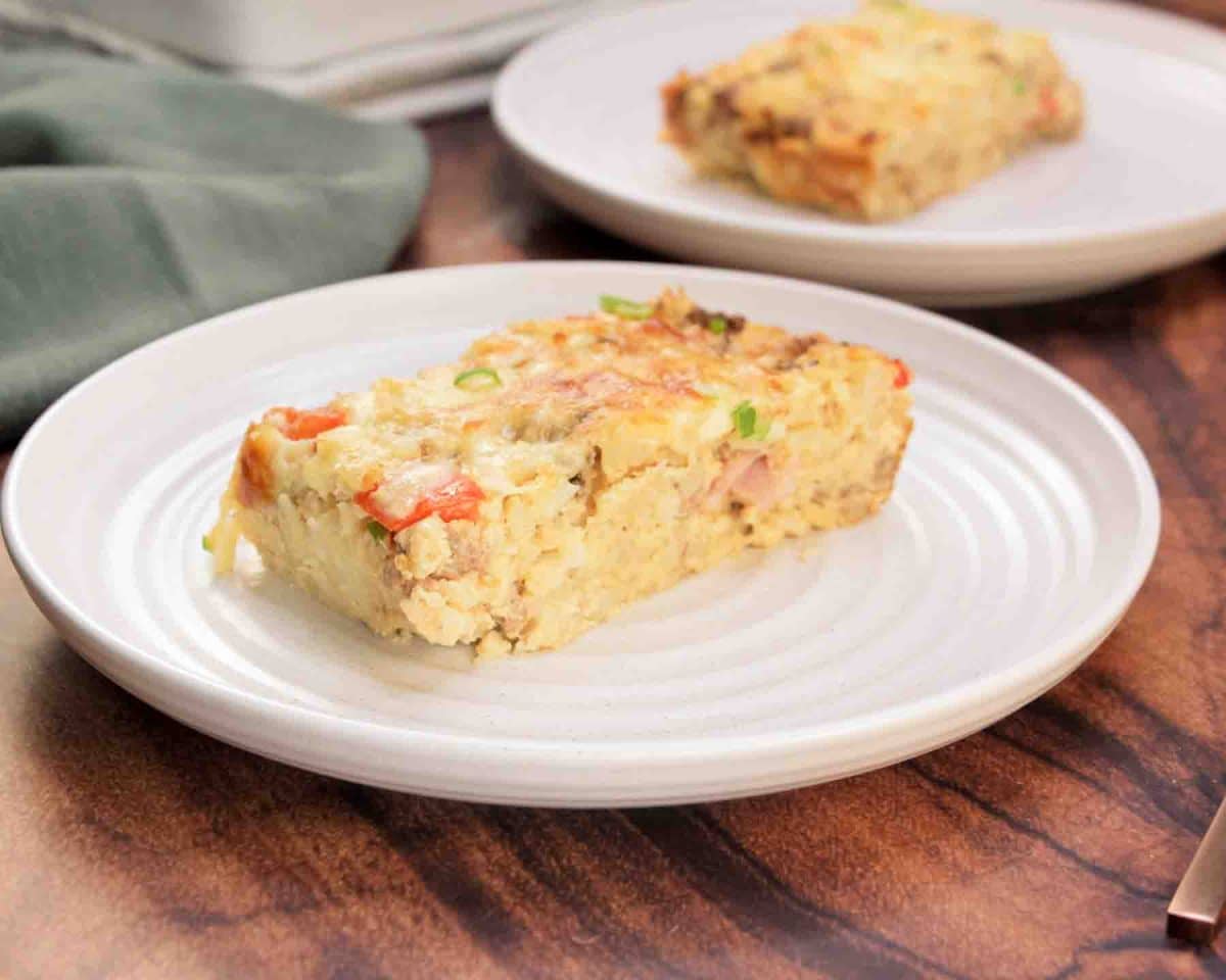 Sideview of a slice of golden brown egg and sausage casserole with melted cheese on a white plate - Hostess At Heart