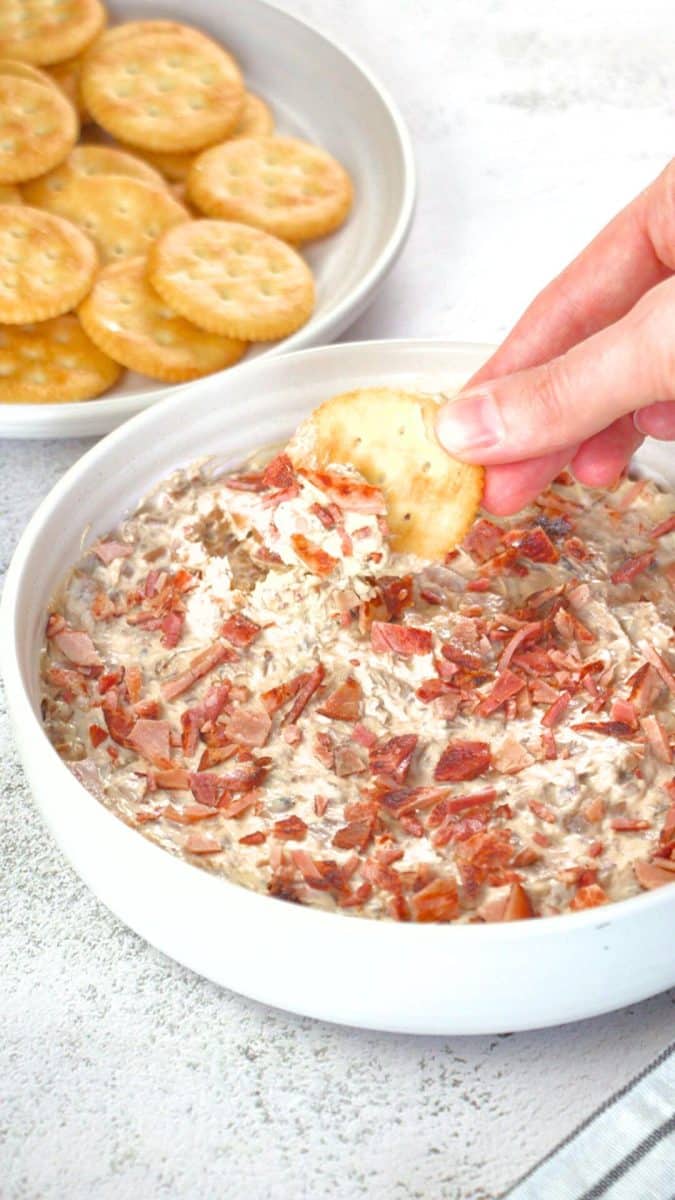A hand dipping a cracker full of caramelized onion bacon dip from a white bowl. Hostess At Heart