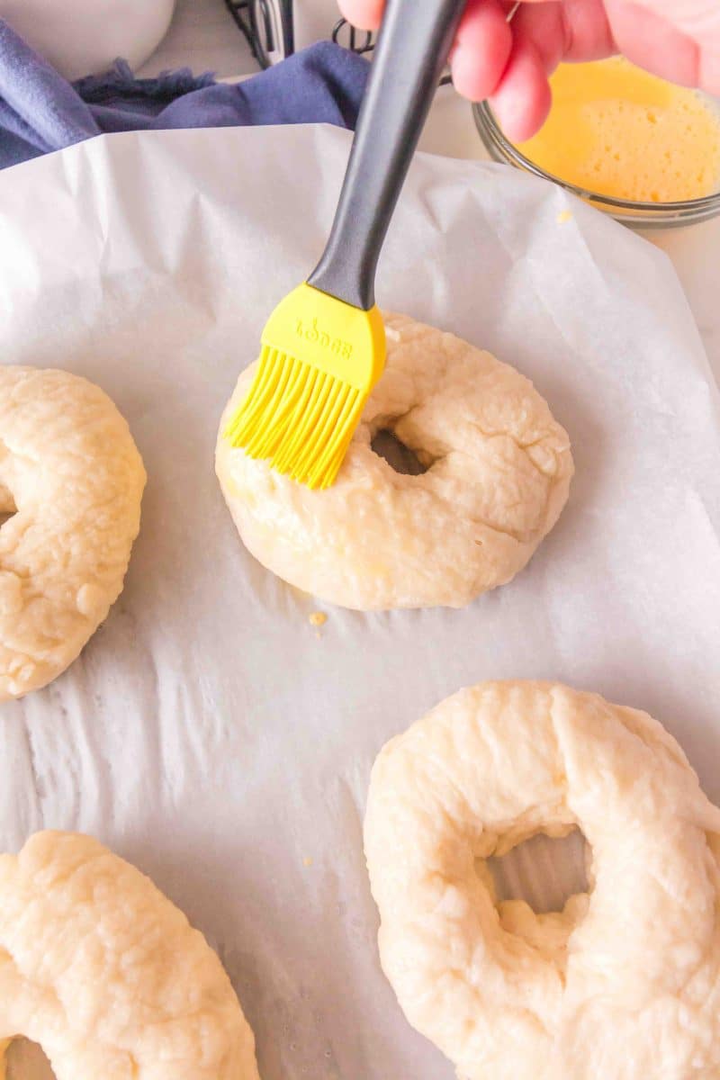 Freshly boiled bagel dough on a parchment paper-covered sheet pan being brushed with egg wash before baking. Hostess At Heart