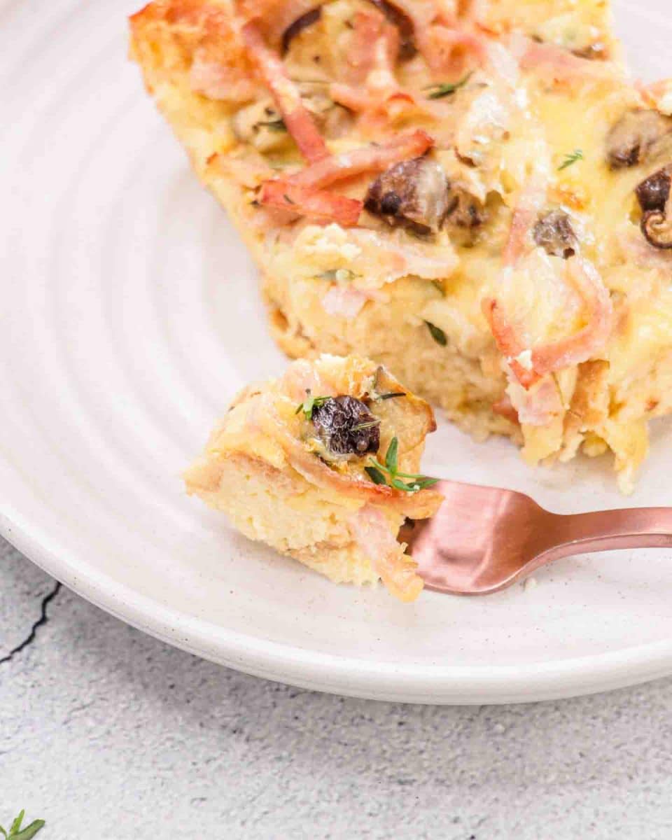 Slice of ham and cheese breakfast strata on white plate with a fork filled with a bite. Hostess At Heart