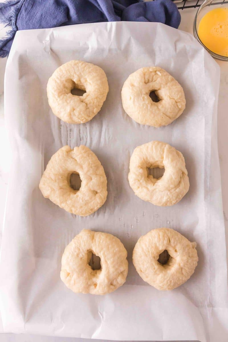 A parchment paper-lined sheet pan filled with boiled homemade yeast bagels before they're baked to a golden brown - Hostess At Heart