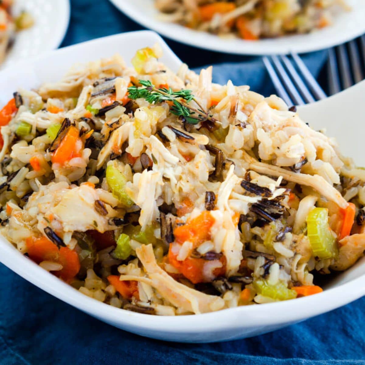 A sideview of a white serving bowl filled with instant pot chicken and wild rice. It's loaded with colorful carrots and celery. Hostess At Heart