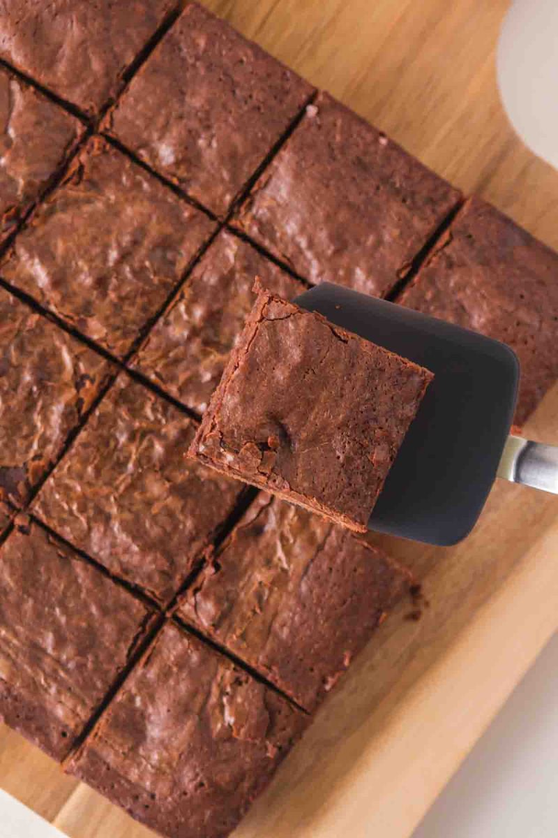 Top view of a brownie on a spatula hovering above the sliced brownies on a wooden cutting board. Hostess At Heart