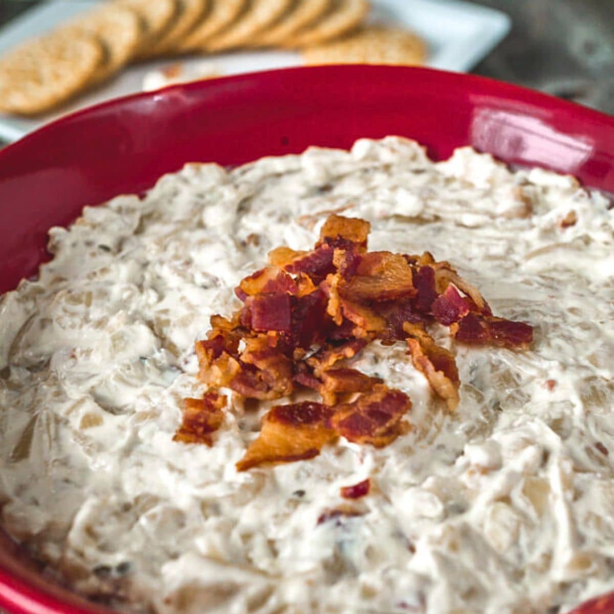 Golden creamy caramelized onion bacon dip topped with crispy bacon pieces in a red serving bowl. Hostess At Heart