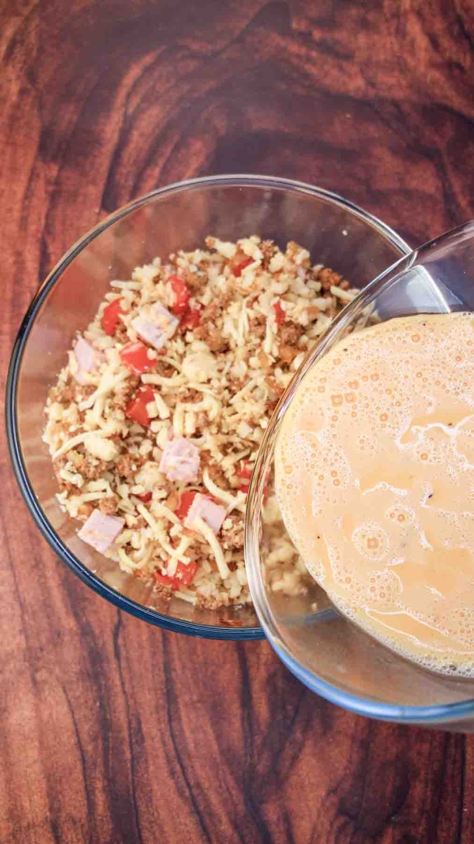 A bowl of an egg mixture being poured over sausage casserole ingredients in glass bowl. Hostess At Heart