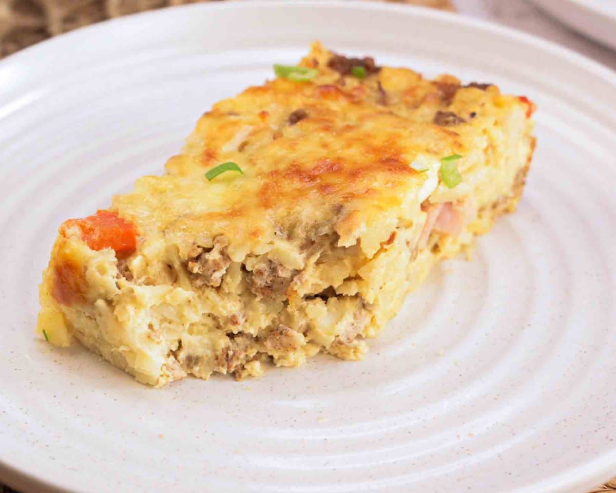 Slice of egg sausage casserole with the front bite removed on white plate showing layers. Hostess At Heart