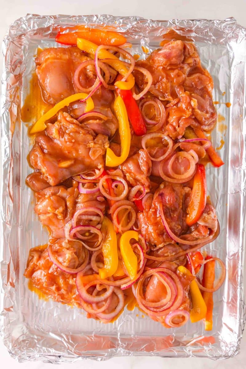 Marinated chicken thighs with sliced bell peppers and onions on foil-lined baking sheet. Hostess At Heart