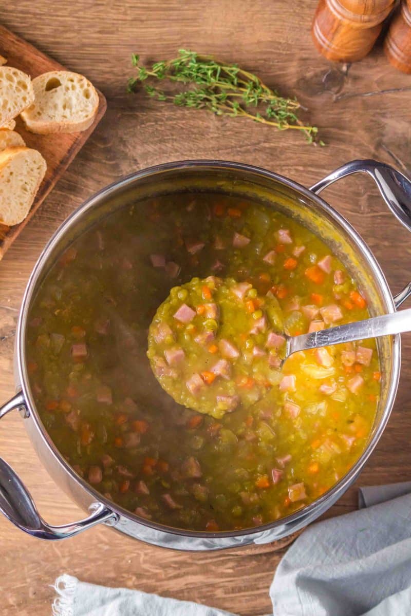 Top down view of a pot of split pea and ham soup with a ladle lifting a serving showing the thick, creamy consistency with a chunk of ham. Hostess At Heart