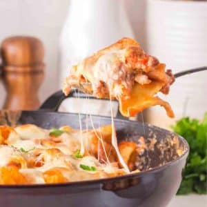 Sideview of a skillet of baked rigatoni pasta with a serving spoon scooping a serving from the skillet. Melted cheese is pulling from the hot Italian sausage pasta bake. Hostess At Heart