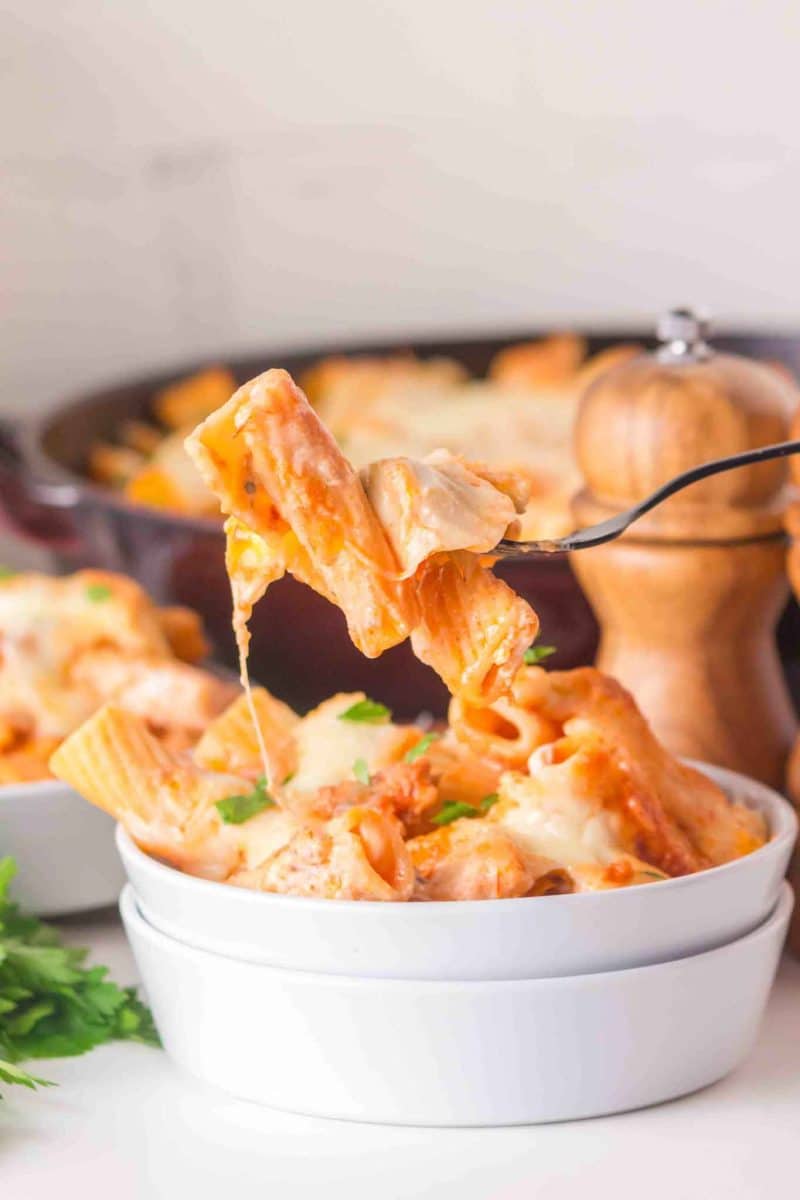 A bowl filled with a baked rigatoni recipe with a fork lifting a stretching melted cheesy bite. Hostess At Heart