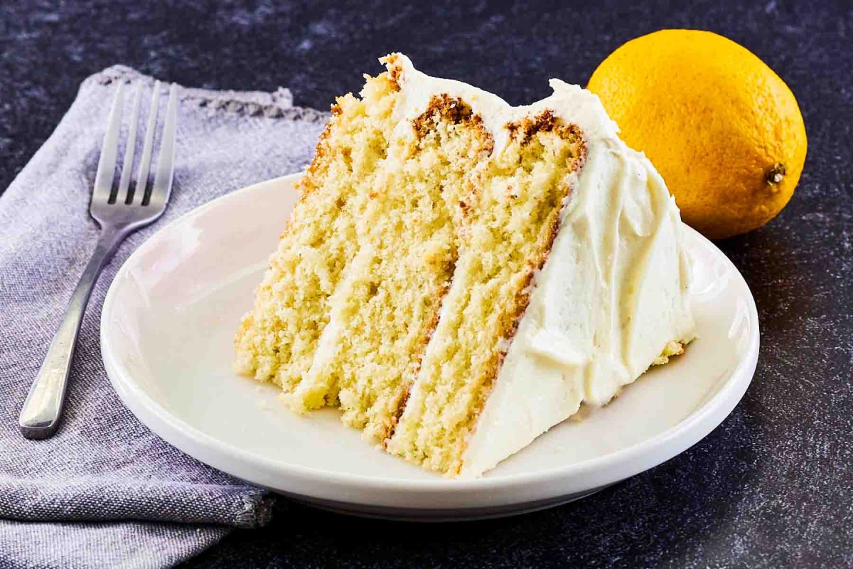 Slice of easy lemon cake with lemon cream cheese frosting on white plate showing moist yellow layers - Hostess At Heart