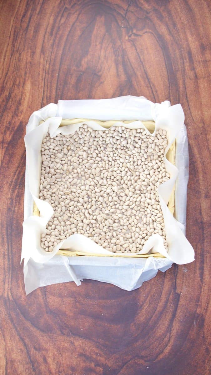 Pie dough fitted in a square parchment paper- lined baking dish and filled with beans for blind baking. Hostess At Heart