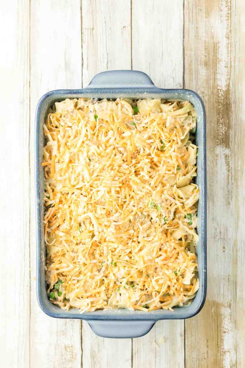 A baking dish filled with tuna casserole ingredients and topped with shredded cheese. Hostess At Heart