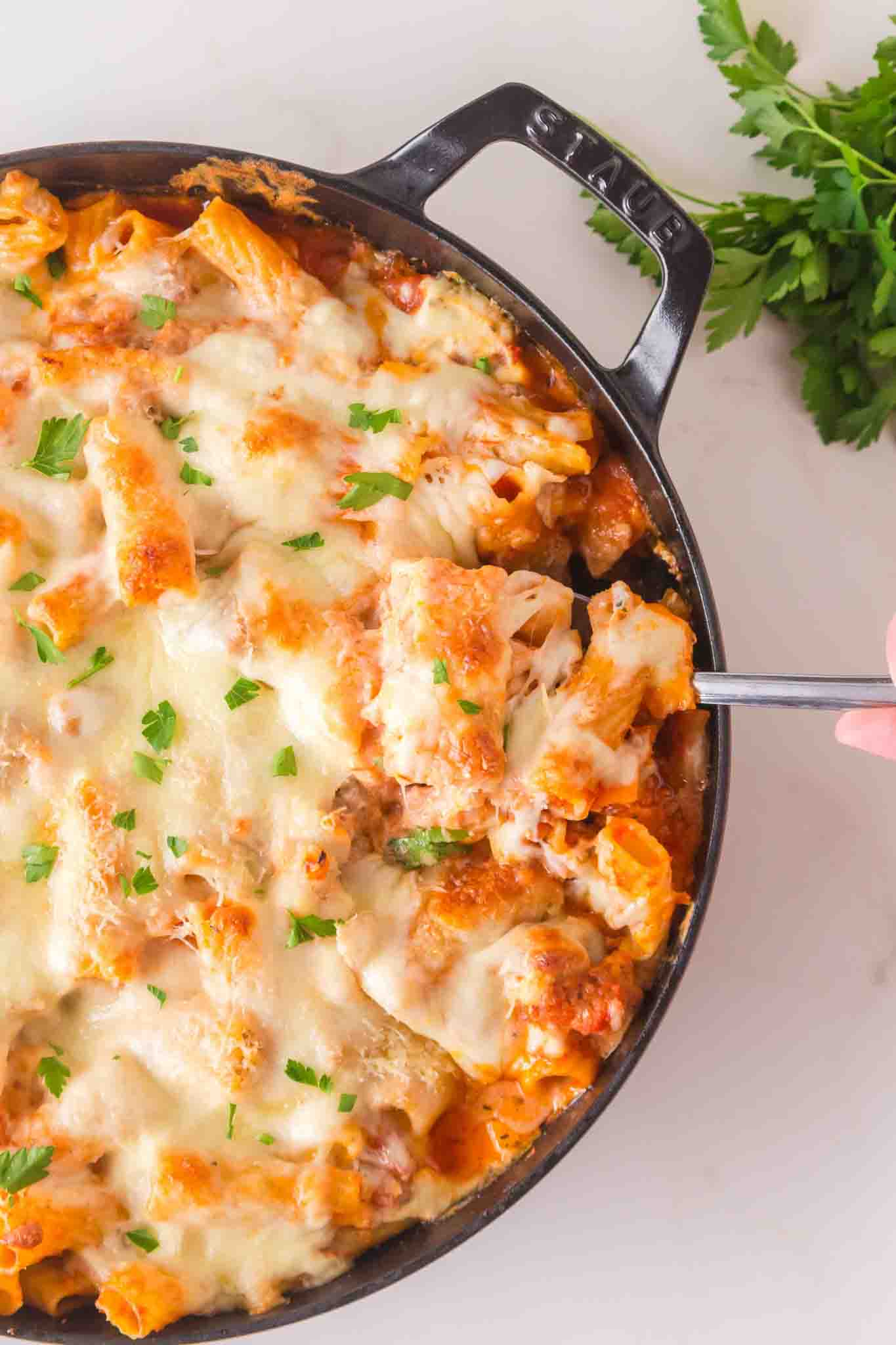 Baked cheesy rigatoni with sausage in cast iron skillet with melted cheese and marinara sauce. A spoon is preparing to dish a serving - Hostess At Heart