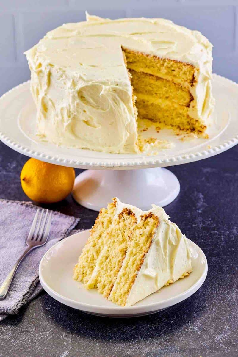 Slice of moist lemon cake with lemon frosting on white plate showing moist yellow layers with the cut layer cake in the back ground. Hostess At Heart