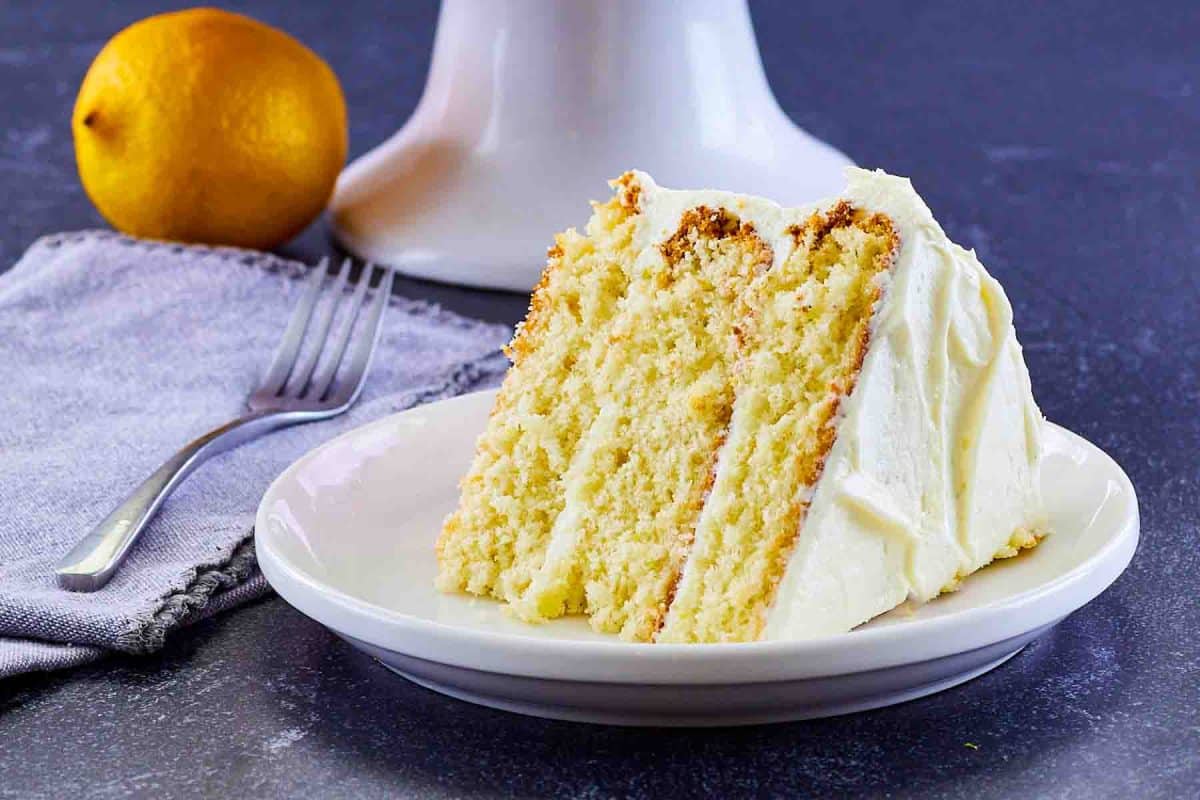 Slice of lemon cake from scratch with lemon cream cheese frosting on white plate showing moist yellow layers. Hostess At Heart