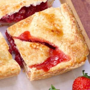 A corner slice of a strawberry slab pie pulled away from the square dessert showing a thick strawberry filling oozing out of a butter crust. Hostess At Heart