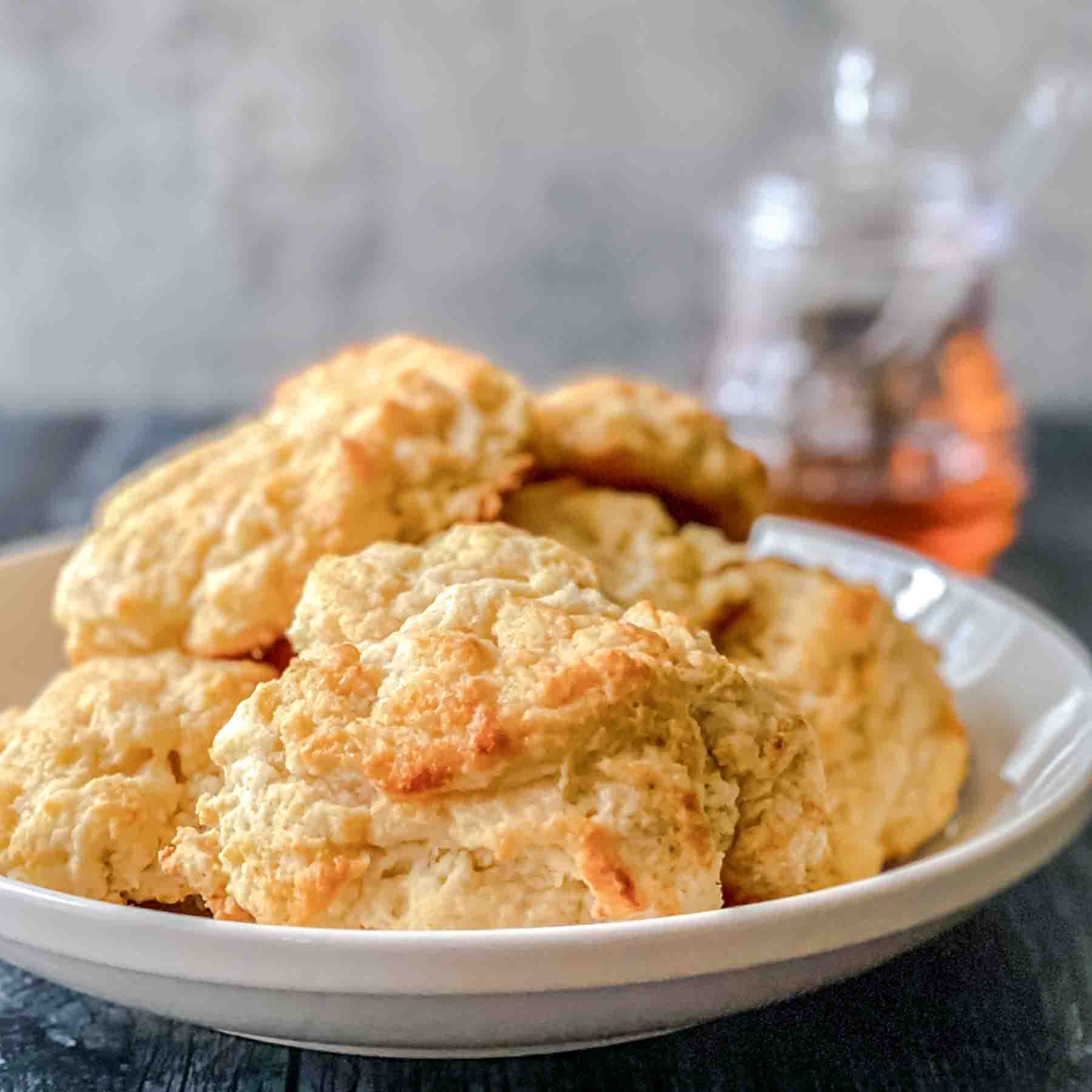 Sideview of a serving bowl filled with golden brown buttermilk drop biscuits topped with honey glaze. Hostess At Heart