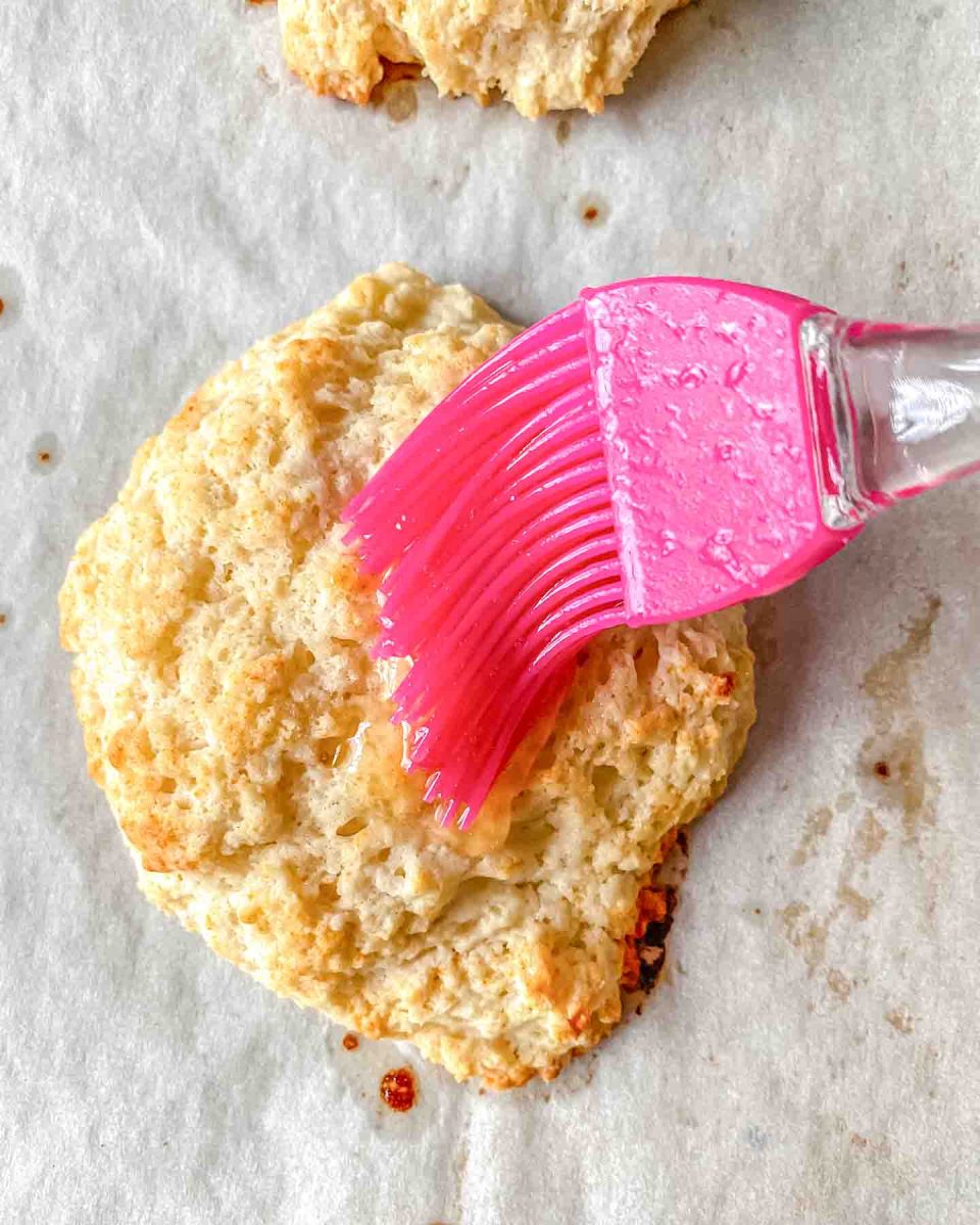 A baked drop biscuit being topped with a honey butter glaze. Hostess At Heart