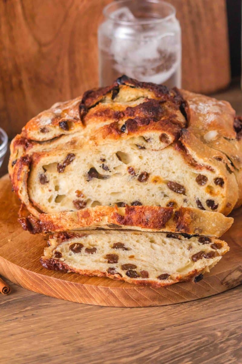 Front view of a loaf of overnight cinnamon raisin sourdough bread sitting on a cutting board with the first two slices cut showing plump raisins and cinnamon swirls throughout a tender crumb. Hostess At Heart