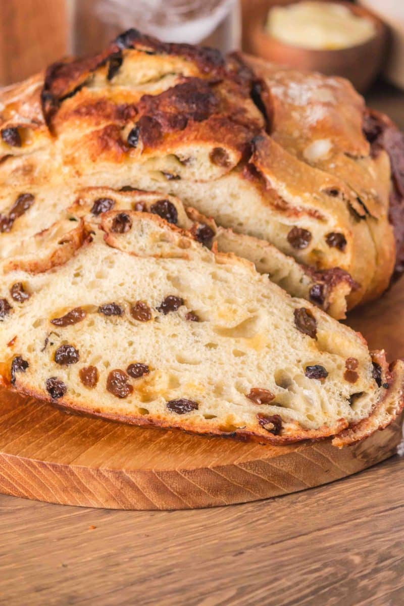 Front view of a loaf of raisin cinnamon sourdough bread sitting on a cutting board with the first two slices cut showing plump raisins and cinnamon swirls throughout a tender crumb. Hostess At Heart