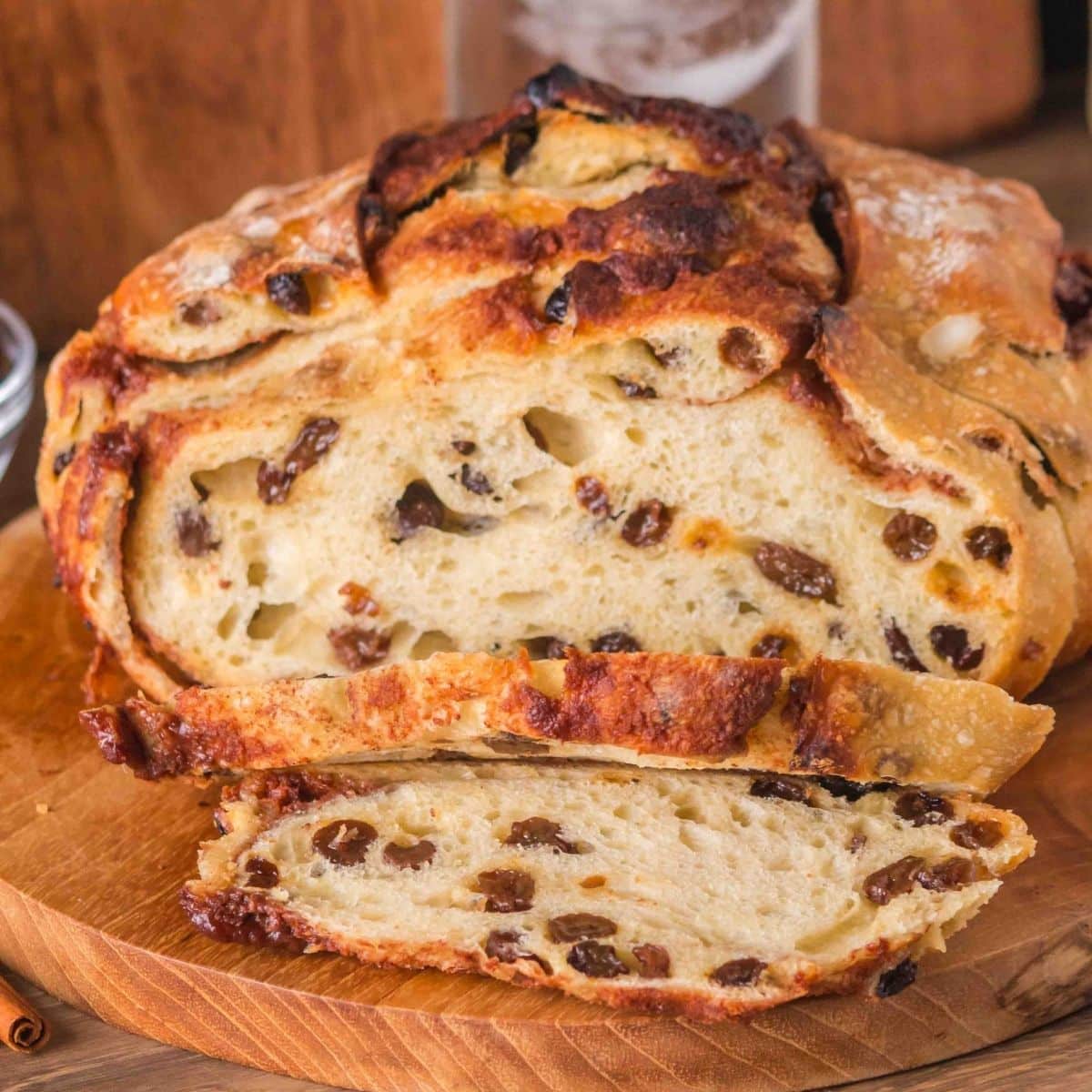 Front view of a loaf of overnight sourdough cinnamon raisin bread sitting on a cutting board with the first two slices cut showing plump raisins and cinnamon swirls throughout a tender crumb. Hostess At Heart
