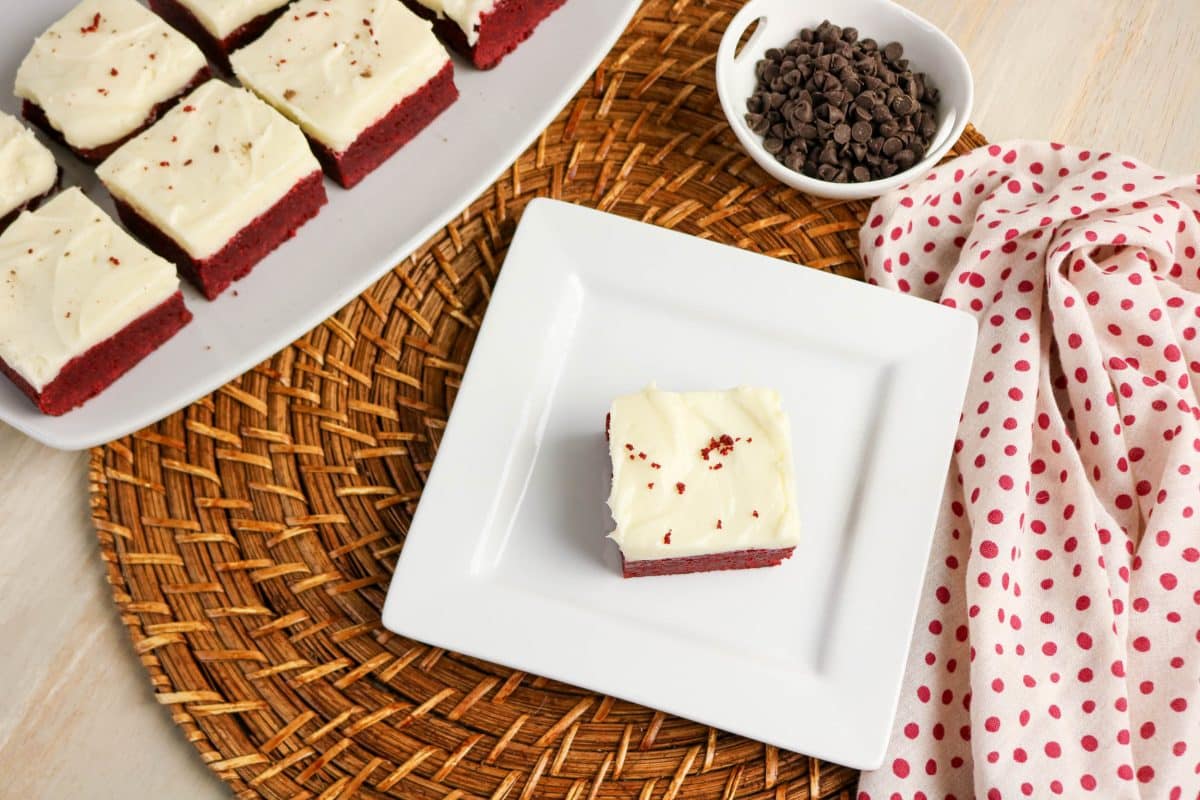 Top down view of a Red velvet brownie on plate beside a serving platter of more fudgy red velvet brownies showing fudgy interior and cream cheese frosting layer - Hostess At Heart