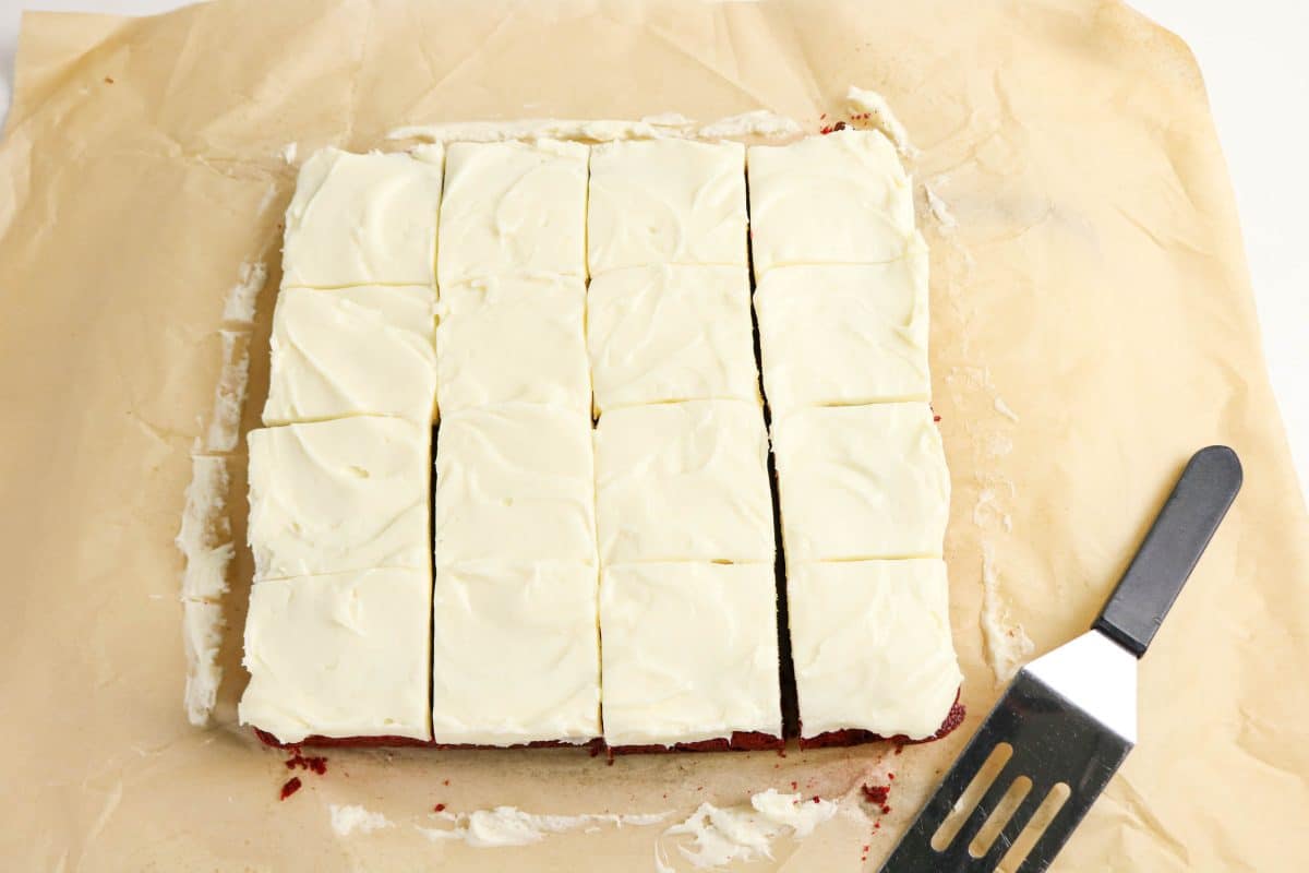 Red velvet brownies topped with cream cheese frosting removed from the baking pan and sliced sitting on parchment paper. Hostess At Heart