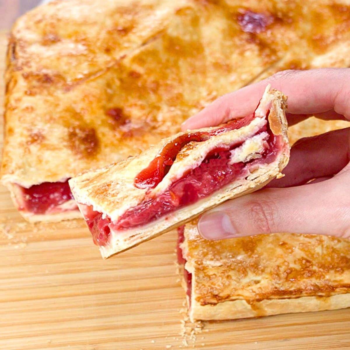 A hand holding a slice of strawberry sheet pie showing thick filling between a double golden brown crust - Hostess At Heart