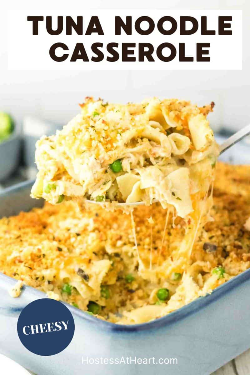 A large spoon lifting a serving of cheesy tuna and noodles casserole from the baking dish showing strings of melted cheese. Hostess At Heart