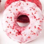 Top down view of a stack of two red velvet donuts. The front one is glazed and sprinkled with red sprinkles for Valentine's Day. The back one is unglazed showing a moist bright red baked donut. Hostess At Heart