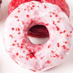 Top down view of a stack of two red velvet donuts. The front one is glazed and sprinkled with red sprinkles for Valentine's Day. The back one is unglazed showing a moist bright red baked donut. Hostess At Heart