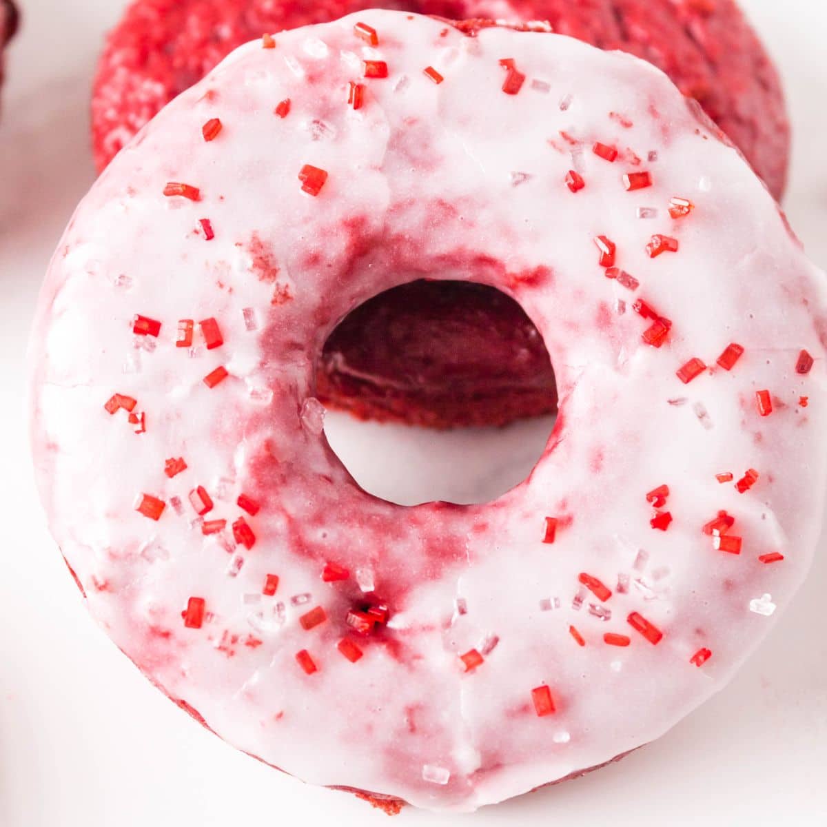 Top down view of a stack of two red velvet donuts. The front one is glazed and sprinkled with red sprinkles for Valentine's Day. The back one is unglazed showing a moist bright red baked donut. Hostess At Heart