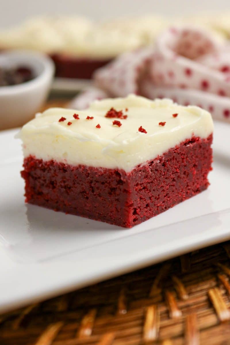 Square red velvet brownie with cream cheese frosting on white plate, showing moist texture and red color. Hostess At Heart