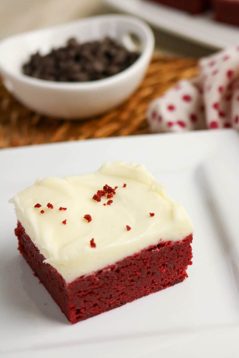 An angled view of a piece of red velvet brownies recipe topped with cream cheese frosting on white plate, showing fudgy texture and a deep red color - Hostess At Heart