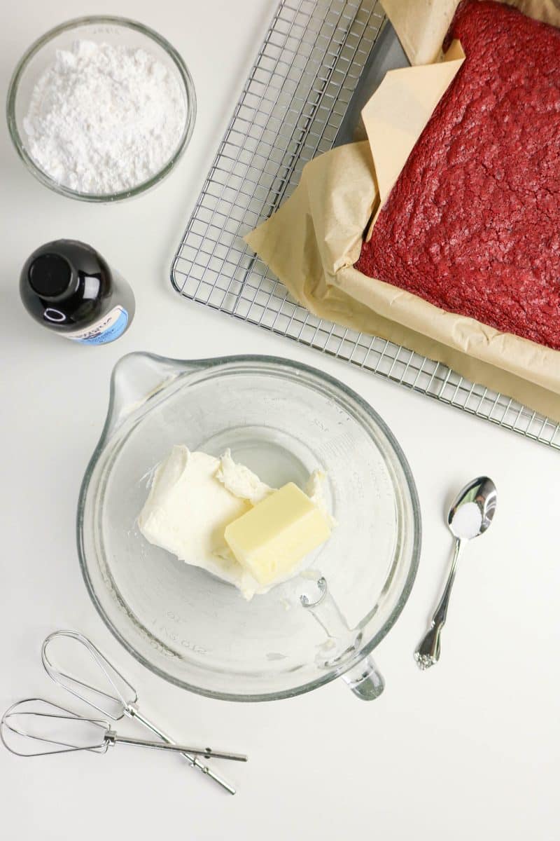 Butter and cream cheese in a mixing bowl before creaming together. Cream cheese ingredients surround the bowl including powdered sugar and vanilla. Hostess At Heart