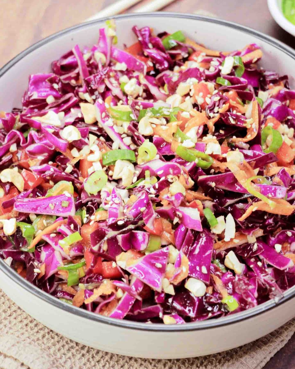 A serving dish filled with an Asian Cabbage Slaw featuring raw purple cabbage, peppers, carrots, in a ginger dressing garnished with cashews - Hostess At Heart
