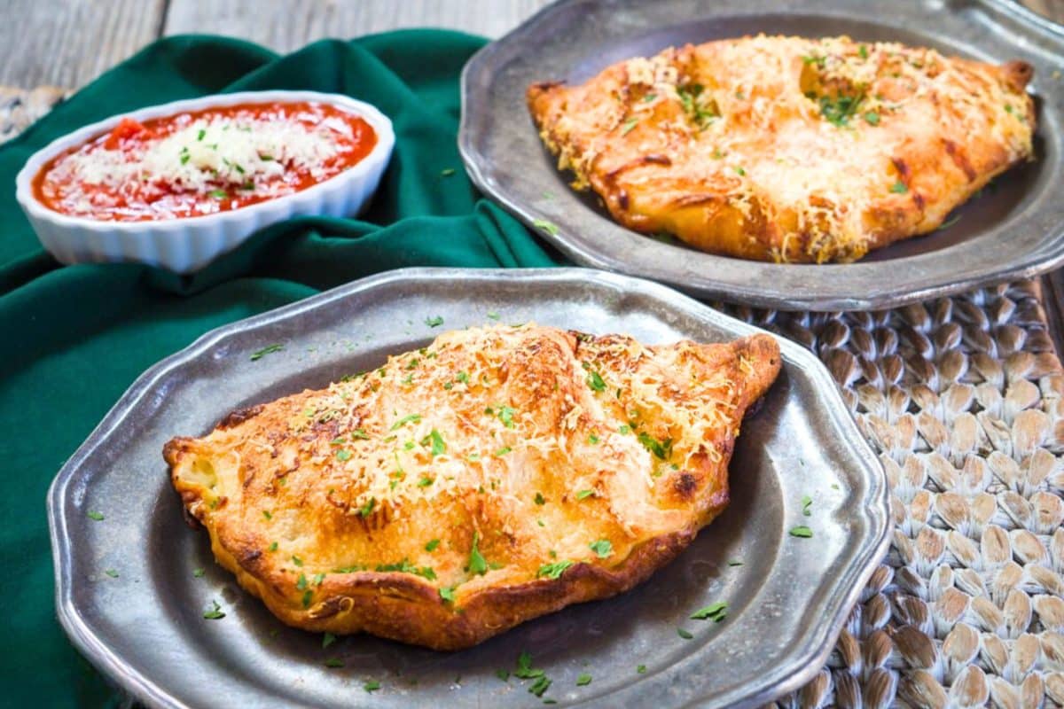Two plates topped with crispy golden brown calzones filled with ham and three cheeses and topped garnished with fresh parmesan. A dish of marinara is ready for dipping. Hostess At Heart