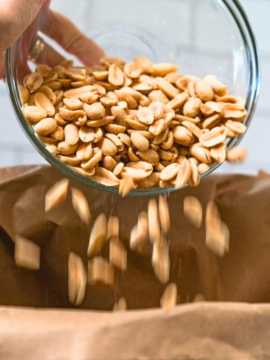 A glass dish filled with salted peanuts being added to a brown paper bag to make a microwave caramel snack mix. Hostess At Heart