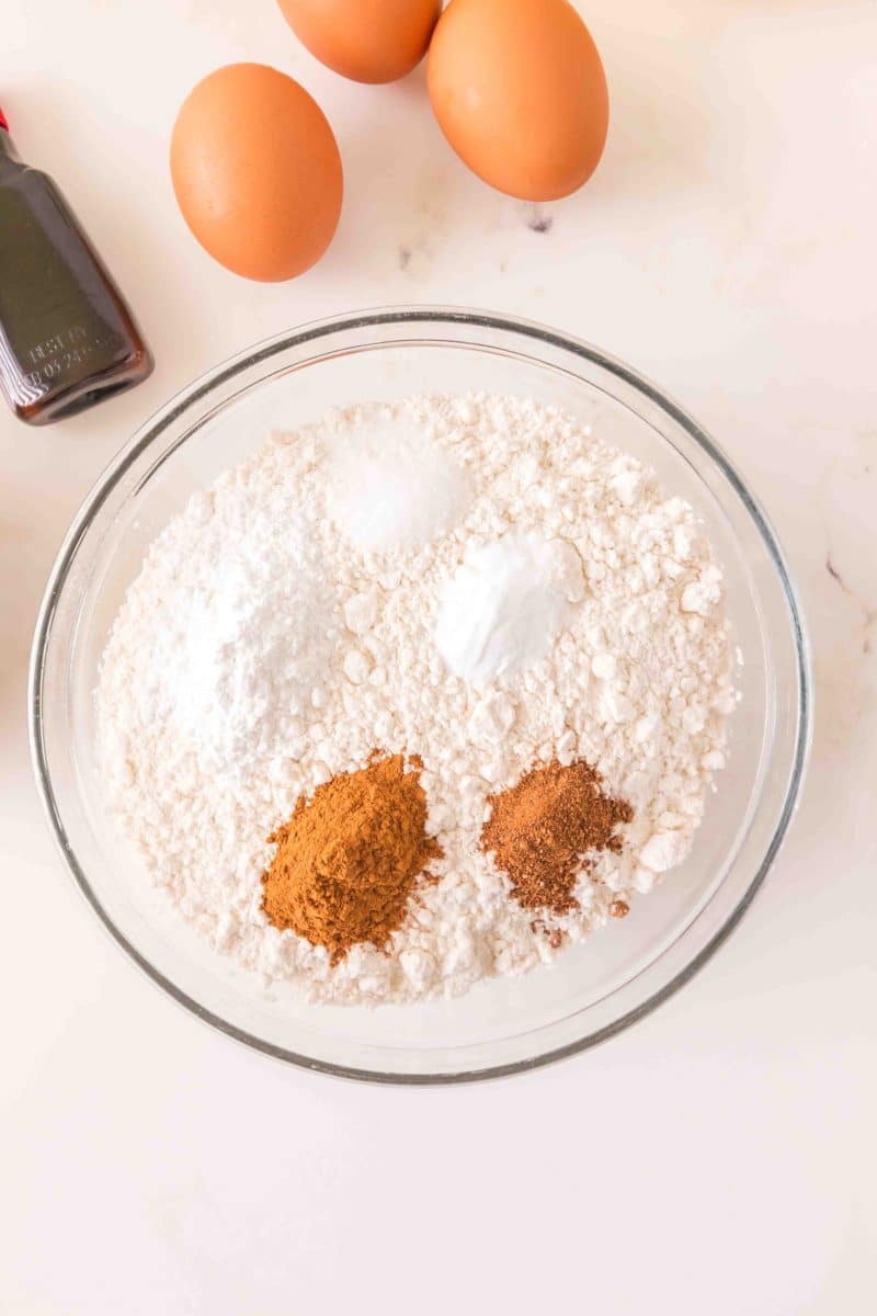 A mixing bowl filled with dry carrot cake recipe ingredients including flour, salt, baking powder, baking soda, cinnamon, and nutmeg. Hostess At Heart