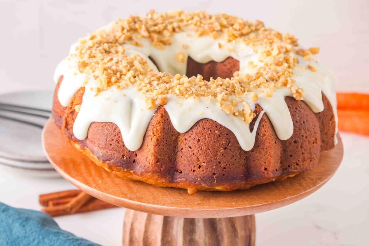 Sideview of a carrot cake with cream cheese glaze garnished with crushed walnuts on a wooden cake stand. Hostess At Heart
