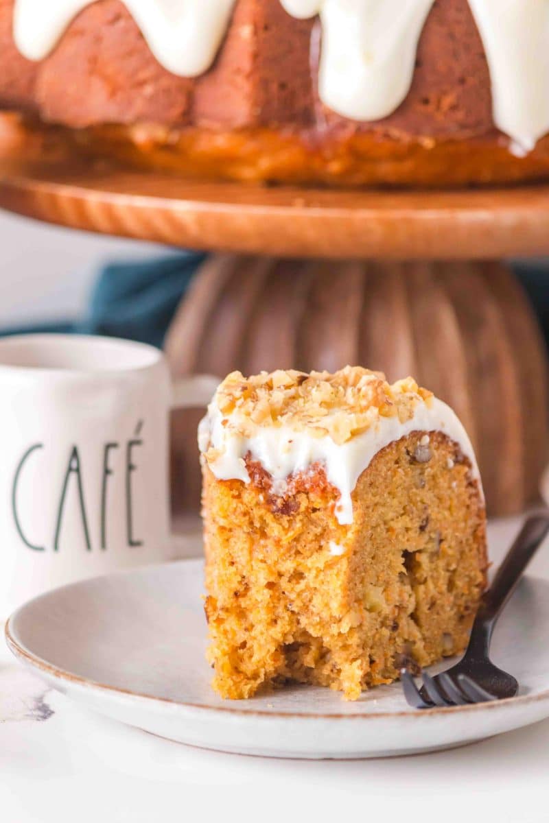 A slice of moist carrot bundt cake with cream cheese glaze on a white plate showing tender texture with visible chopped walnuts. Hostess At Heart
