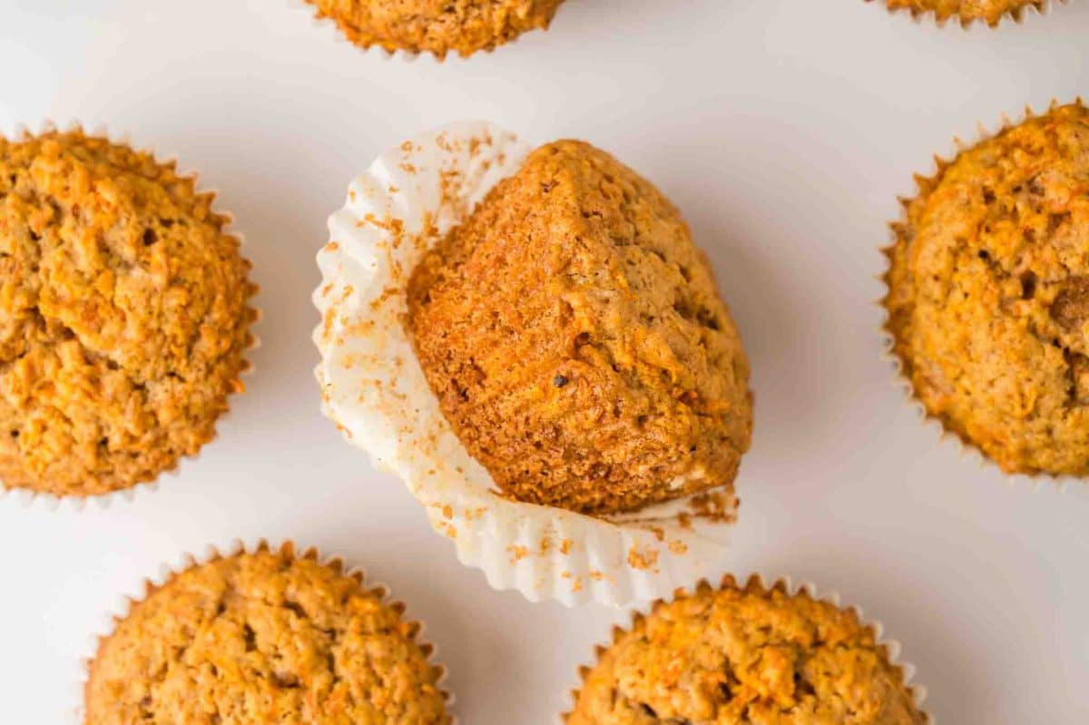 Several carrot cake muffins sitting on a counter with the middle one sitting on it's side over the muffin paper showing a moist golden brown muffin. Hostess At Heart