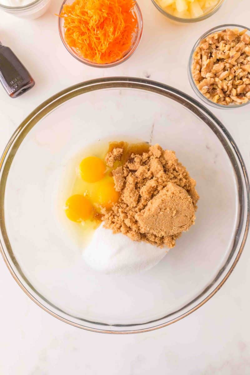 A mixing bowl filled with wet carrot cake ingredients ready for creaming including white and brown sugar and eggs. Hostess At Heart