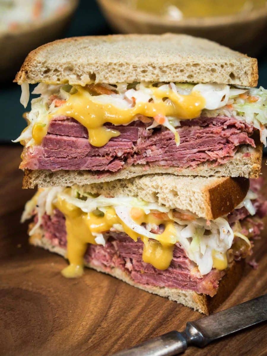 Two halves of a corned beef sandwich stacked on each other showing layers of moist corned beef, crunchy cabbage slaw, and a rich mustard sauce on homemade rye bread. Hostess At Heart