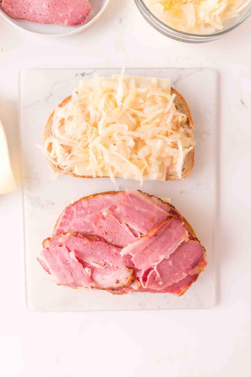 Two slices of rye bread topped with thousand island dressing, Swiss Cheese, Sauerkraut, and slices of Corned Beef showing how to make a reuben sandwich. Hostess At Heart