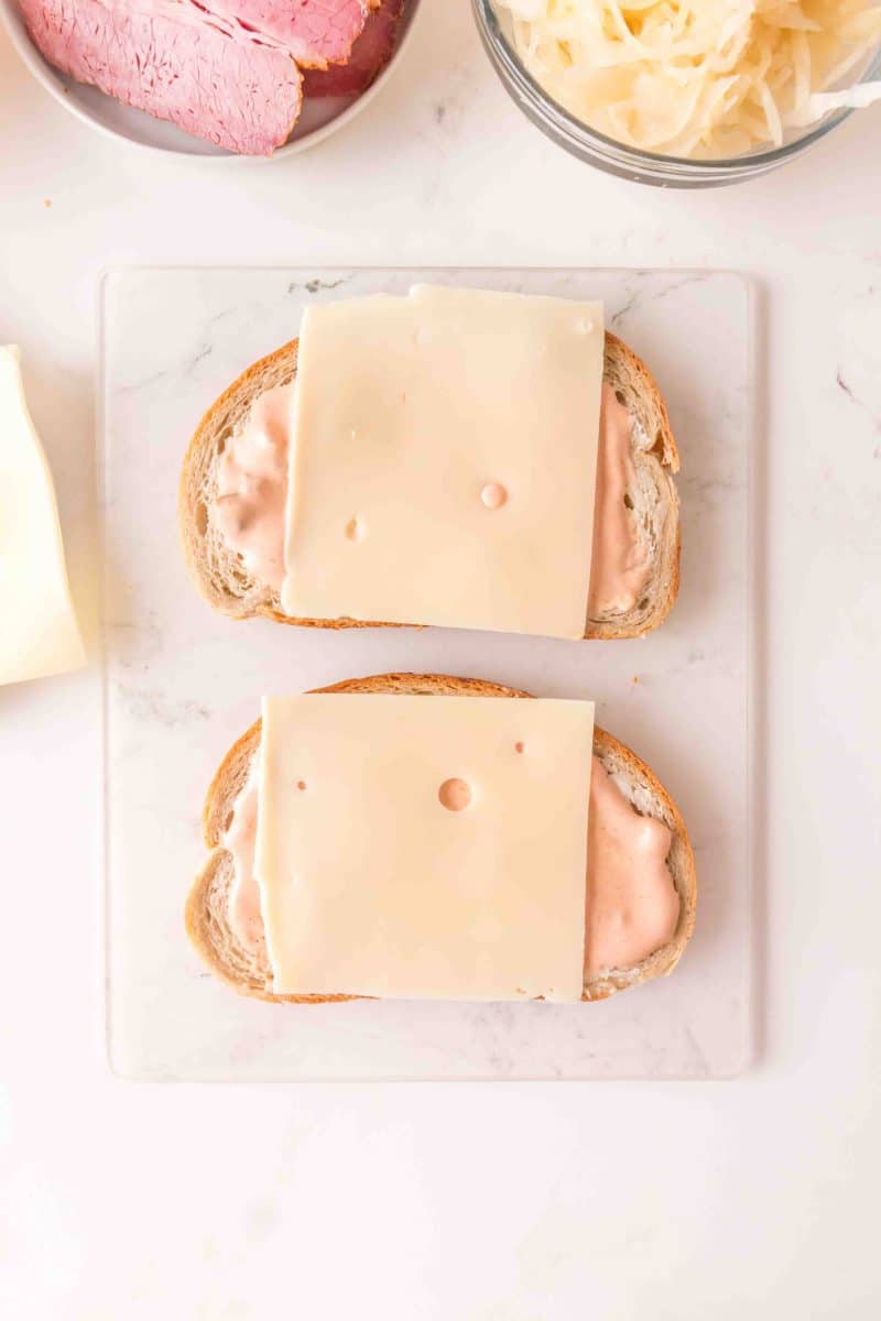 Sliced Swiss cheese placed on rye bread slathered with thousand island dressing demonstrating how to make a Reuben Sandwich recipe. Hostess At Heart