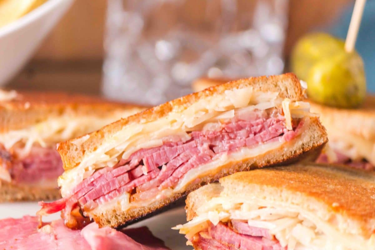 A half of a Reuben sandwich propped up on cut sandwiches showing a thick layer of sliced corned beef, melted Swiss cheese, sauerkraut, and creamy thousand island dressing between two slices of golden brown grilled rye bread. Hostess At Heart