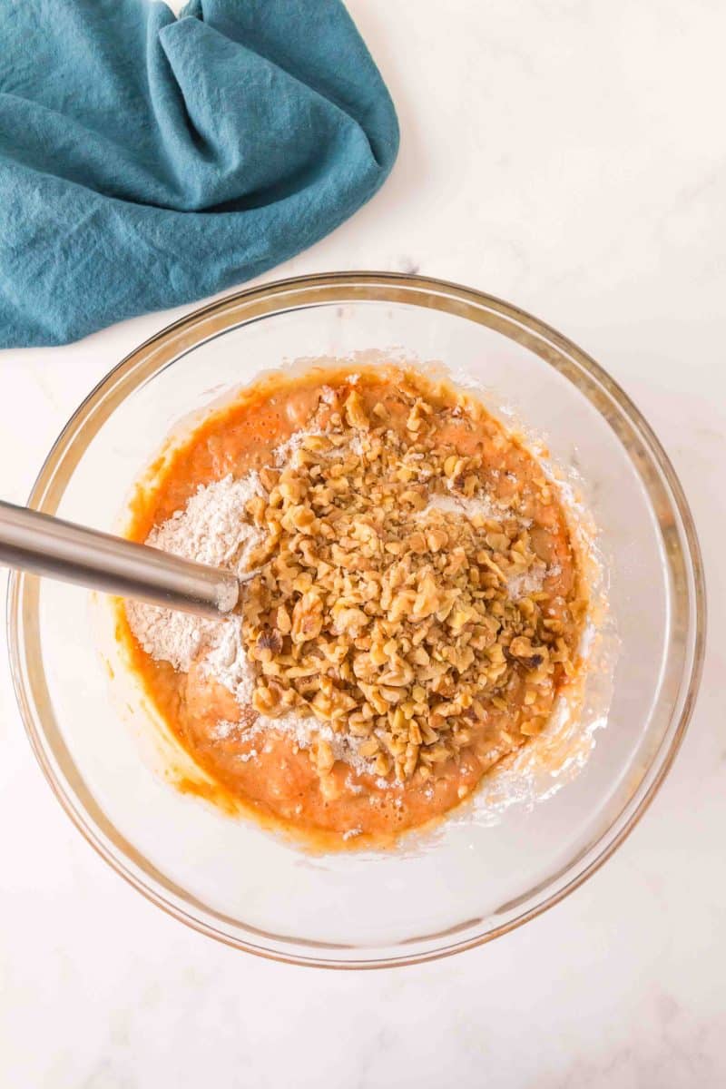 Top down view of crushed nuts added to a thick carrot bundt cake batter in a mixing bowl. Hostess At Hearts